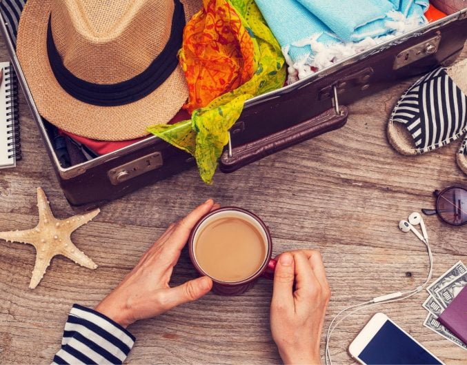 Suitecase filled with items for a vacation. Woman holding coffee cups in her hands.